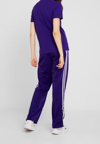 Lila atletisk outfit bestående av en tight T-shirt och lösa byxor med vita ränder längs sidorna. Mönsterfritt tyg med en jämn textur.