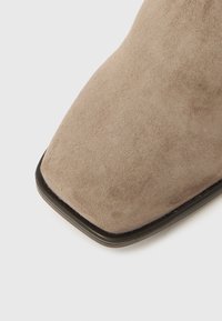 Beige semsket ankelstøvel med firkantet tå, med en glatt tekstur og en kontrasterende svart såle. Enkel design uten pynt.