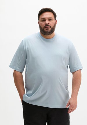 Homme avec une barbe portant un T-shirt bleu clair à manches courtes et un pantalon noir, debout avec une main dans la poche sur fond blanc.