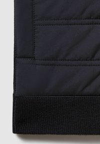 Colț de jachetă puffer neagră, cu o manșetă texturată din tricot cu nervuri și un model matlasat orizontal, realizat dintr-un material ușor și neted.