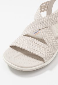 Helle beige Sandalen mit überkreuzten, gewebten Stoffriemen, strukturiertem Fußbett und einer soliden, cremeweißen Sohle mit kleinen Perforationen.
