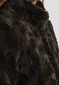 Fuskpälsjacka i mörkbrunt med en mjuk, texturerad yta och hög krage. Tjockt, yvigt material med ojämn lugg ger djup.