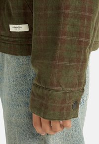 Zielona kurtka z korduroju w kratkę, z czarnym guzikiem i etykietą z napisem "ELEMENT CO. SINCE 1992". Noszona na niebieskim dżinsie.