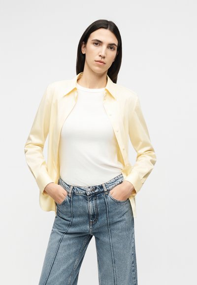 Γυναίκα με ίσια σκούρα μαλλιά φοράει ένα ανοιχτό κίτρινο πουκάμισο με κουμπιά πάνω από λευκό τοπ, ψηλόμεσα μπλε τζιν παντελόνια, με τα χέρια στις τσέπες, ουδέτερο φόντο.