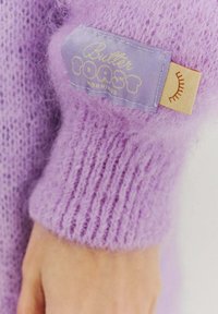 Fialový pletený svetor s chlpatou textúrou. Má svetlofialovú štítok s nápisom "Butter Toast" a oranžový slnečný motív.