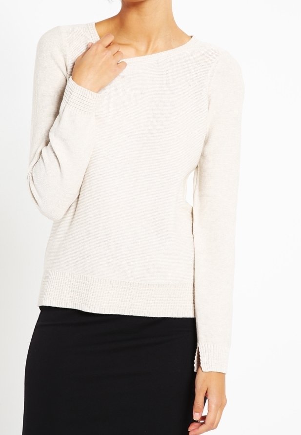 Frau in einem langärmligen beigen Strickpullover mit strukturierten Bündchen und Saum, kombiniert mit einem schwarzen Rock, posiert mit einer Hand in der Nähe des Schlüsselbeins.