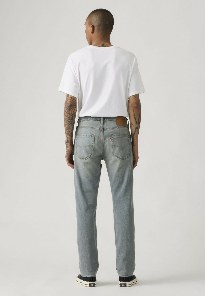 Homme tatoué portant un t-shirt blanc, un jean bleu clair et des baskets noires, debout de dos devant un fond gris uni.