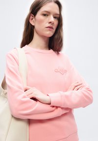 Νεαρή γυναίκα με μακριά καστανά μαλλιά, φορώντας ροζ φούτερ και εκρού τσάντα ώμου, με σταυρωμένα τα χέρια, μπροστά σε φωτεινό μονόχρωμο φόντο.