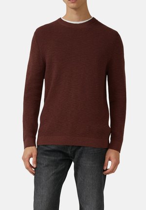 Mann, der einen dunkelroten strukturierten Strickpullover über einem weißen Rundhals-Shirt trägt, kombiniert mit verblichenen schwarzen Jeans, vor einem schlichten Hintergrund.