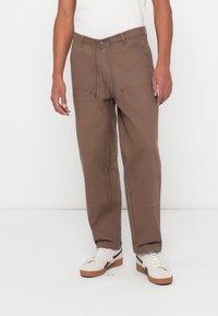 Pantaloni intrecciati marroni con una vestibilità rilassata, dotati di cordino regolabile in vita e due tasche anteriori; abbinati a sneaker bianche.