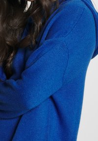 Blå stickad tröja med avslappnad passform, med detaljerad textur och huva. Ärmarna är något sänkta, vilket betonar komfort.