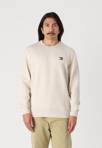 Lys beige langermet genser med rund hals, ribbede mansjetter, og en liten flerfarget merke-logo på venstre bryst.