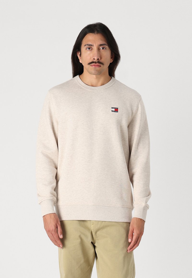 Lys beige langermet genser med rund hals, ribbede mansjetter, og en liten flerfarget merke-logo på venstre bryst.