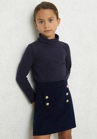 Námornícky rolák s dlhým rukávom skombinovaný s tmavomodrou sukňou so zlatými gombíkovými akcentmi a bočnými vreckami. Mäkko textúrovaná látka.