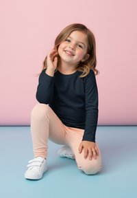 Granatowa bluzka z długim rękawem i okrągłym dekoltem, pastelowo-różowe legginsy oraz białe trampki. Dziecko siedzi, trzymając rękę na włosach, uśmiecha się.