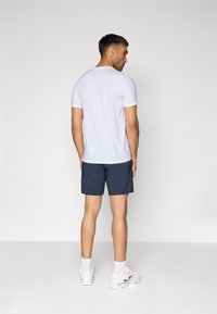 Camiseta atlética de mangas curtas e brancas combinada com calções de cor azul-marinho, vista por trás. Apresenta linhas limpas e uma textura suave, usada com ténis brancos.