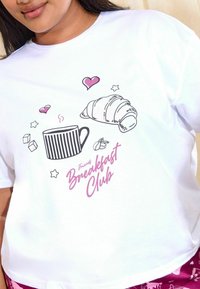 Valge T-särk, millel on graafika triibulisest tassist, kroissantist, südametest ja roosast tekstist "Friends Breakfast Club".