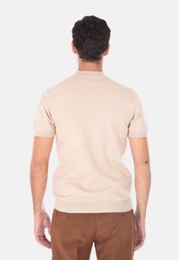 Uomo in piedi con le spalle rivolte, indossa un maglione beige a maniche corte e pantaloni marroni, contro uno sfondo chiaro e uniforme.