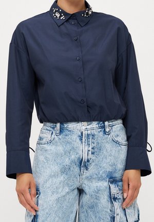 Donkerblauw overhemd met knopen en een met juwelen versierde kraag, gecombineerd met high-waisted acid-wash denim cargobroek, handen ontspannen langs het lichaam.