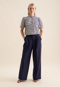 Pantaloni larghi blu abbinati a una blusa bianca e blu con fantasia, caratterizzata da un colletto alto e maniche a sbuffo, con un dettaglio a chiave centrale.