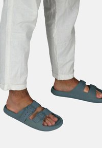 Sandales en caoutchouc grises à la forme arrondie, dotées de deux sangles réglables avec boucles, portées avec un pantalon blanc légèrement fuselé.