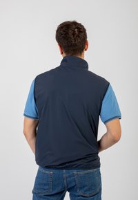 Gilet senza maniche blu navy con colletto alto, realizzato in materiale liscio. Indossato sopra una polo azzurro chiaro con dettagli blu scuro.