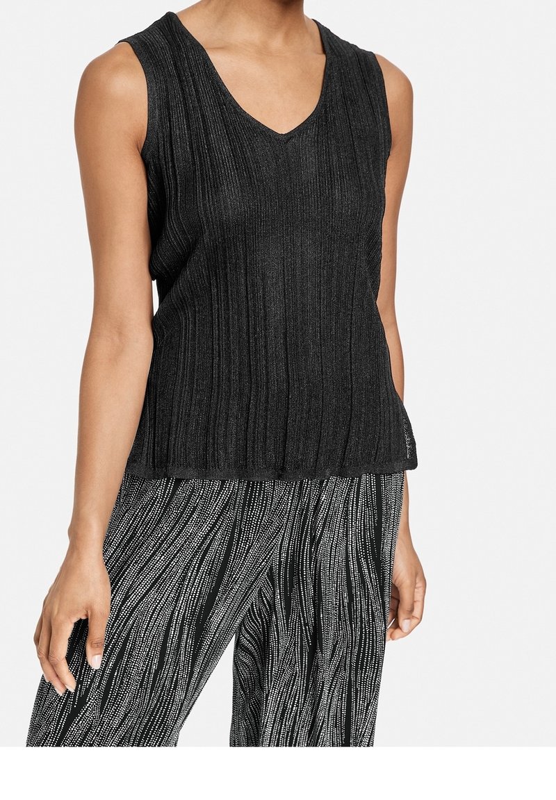 Femme portant un haut sans manches noir côtelé à col en V et un pantalon à motifs noir et blanc avec des lignes ondulées ponctuées.