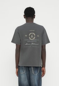 Γκρι βαμβακερό μπλουζάκι με κοντά μανίκια και λαιμόκοψη γύρω από το λαιμό. Στο πίσω μέρος εμφανίζεται ένα γραφικό σχέδιο με κείμενο σε μπεζ και μαύρες λεπτομέρειες.