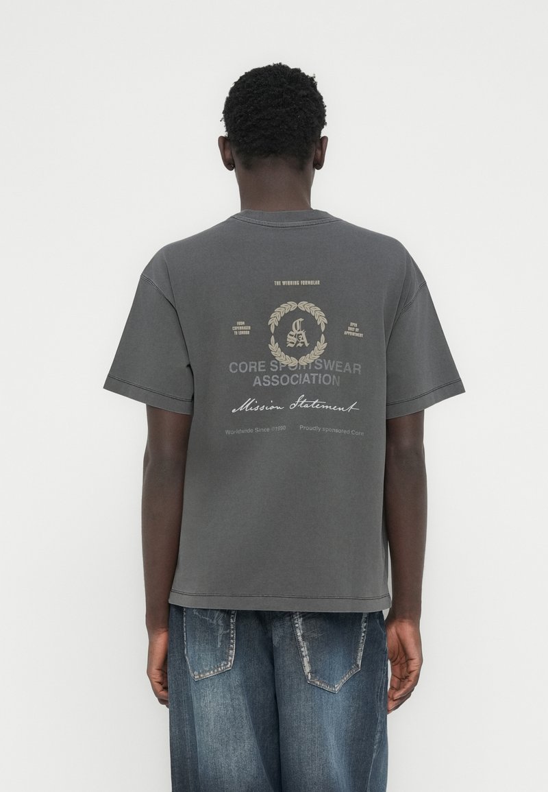 Γκρι βαμβακερό μπλουζάκι με κοντά μανίκια και λαιμόκοψη γύρω από το λαιμό. Στο πίσω μέρος εμφανίζεται ένα γραφικό σχέδιο με κείμενο σε μπεζ και μαύρες λεπτομέρειες.