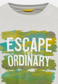 T-shirt blanc avec le texte "ESCAPE THE ORDINARY" sur un motif abstrait de coups de pinceau verts et jaunes à l'avant.