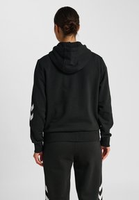 Osoba z włosami w kok klasycznym, ubrana w czarną bluzę z kapturem oraz pasujące czarne spodnie z białymi wzorami w chevron na rękawach i bokach spodni, widok z tyłu.