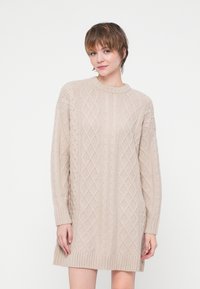 Rochie tricotată bej cu un design cu fire înfășurate, guler rotund, mâneci lungi și tiv cu nervuri. Textură moale și croială lejeră.