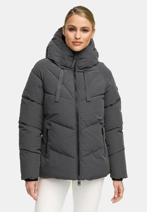 Vrouw in een donkergrijze gewatteerde winterjas met een hoge kraag en rits aan de voorkant, gecombineerd met witte broeken, staand tegen een effen achtergrond.