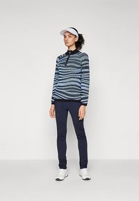 Ženska stoji oblečena v dolgo majico s plavimi in črnimi črtami, mornarsko modre legice, bele superge in belo visor kapo, proti enotnemu ozadju.