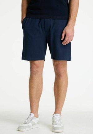 Moški, ki nosi temno modre kratke hlače, bele superge z sivimi poudarki, stoji ob belem ozadju, roke sproščeno ob straneh.