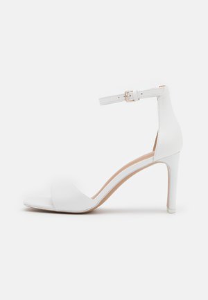 Hvid højhælet sandal med ankelrem, åben tå, glat syntetisk materiale og slank hæl. Simpelt design med minimale detaljer.