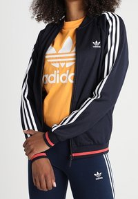 Námořnická sportovní bunda na zip s bílými pruhy podél rukávů, červenými akcenty na lemu, nošená přes žluté tričko s bílým logem.