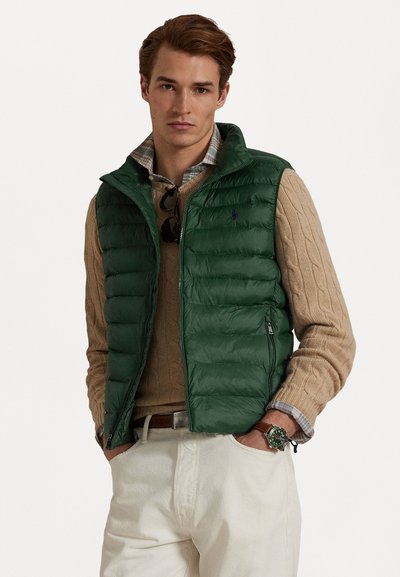 Grøn puffer vest med lynlås, båret over en beige strikket sweater. Har en høj krave og et lille logo på brystet.