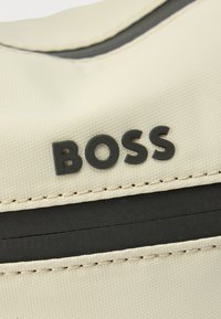 Beežist kangast kott, millel on sile tekstuur, must rõhutusriba ja kõrgendatud must "BOSS" kiri. Minimalistlik disain õmmeldud servadega.