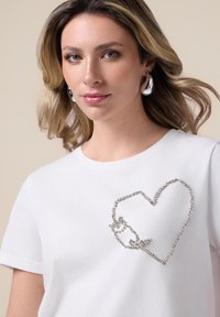 Camiseta de algodón blanca con un diseño de corazón hecho de rhinestones plateados en la parte delantera. Mangas cortas y un clásico escote redondo.