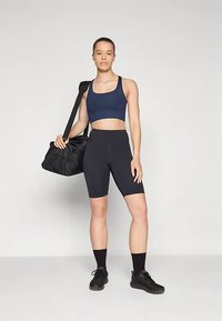 Temno modra športna kratka majica in črne pajkice z visokim pasom. Model drži črno torbo za športno opremo. Črni športni copati dopolnjujejo oblačilo. Nevtralen ozadje.