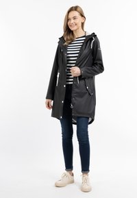 Giacca nera impermeabile con cappuccio, vita regolabile con coulisse e tasche frontali. Abbinata a una camicia a righe, jeans scuri e sneakers chiare.