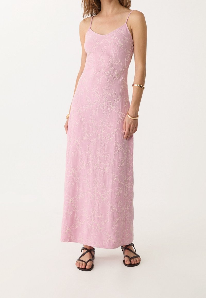 Femme portant une robe maxi rose sans manches avec broderie florale, bracelets dorés, et sandales noires à brides, debout devant un fond uni.