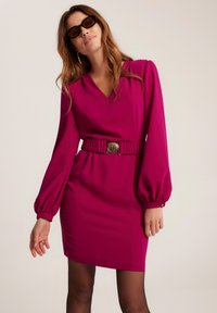 Fuksjowa sukienka z długimi, obszernymi rękawami i dekoltem w szpic. Posiada marszczoną talię i złotą klamrę paska. Gładka tekstura materiału.
