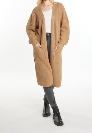 Femme blonde portant un long cardigan en maille couleur camel sur un haut blanc, un jean gris et des bottines noires, debout les mains dans les poches.