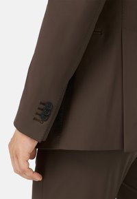 Ruskea puku takki, jossa on sileä pinta, istuvat hihat ja neljä mustaa nappia tikkauskoristeilla.