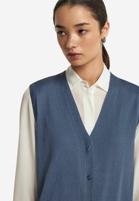 Ärmlös blå stickad väst med V-hals, med ribbstruktur och tre mörka knappar. Bärs över en vit skjorta med knappar.