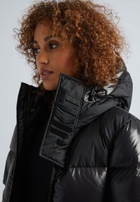 Chaqueta de plumas negra con un cuello alto y el logotipo bordado "JKIT" en el lado izquierdo; presenta un acabado brillante y una textura suave.