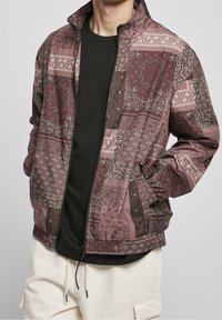 Mörkröd och beige bomberjacka med paisleymönster, dragkedja, elastisk nederkant och fickor. Bärs över en svart skjorta. Slät textur.