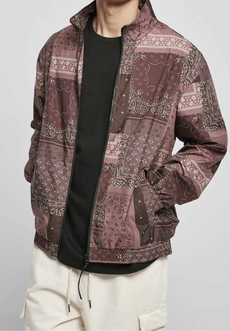 Mörkröd och beige bomberjacka med paisleymönster, dragkedja, elastisk nederkant och fickor. Bärs över en svart skjorta. Slät textur.
